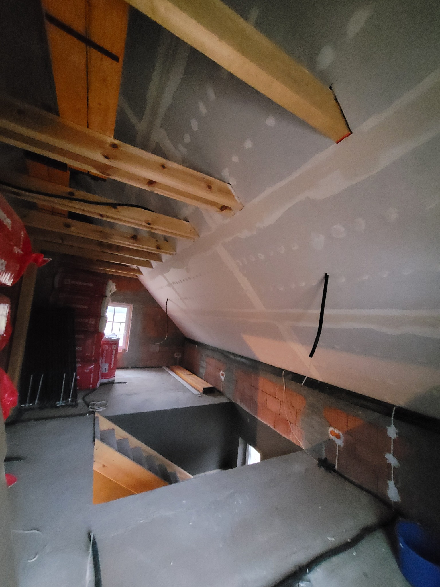 Poddasze w trakcie wykańczania: widoczne krokwie, częściowo ocieplone ściany z płyt gipsowo-kartonowych, ceglana ściana z instalacją elektryczną, okno w tle i izolacja termiczna w czerwonych...