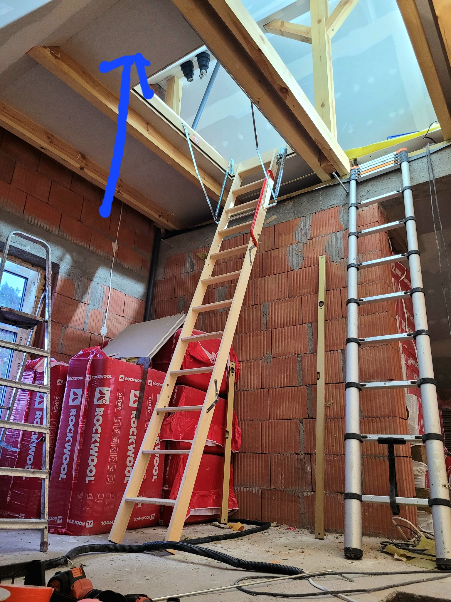 Montaż schodów strychowych w surowym stanie, widoczne ocieplenie wełną mineralną Rockwool, ceglana ściana, drabina aluminiowa i poziomica.