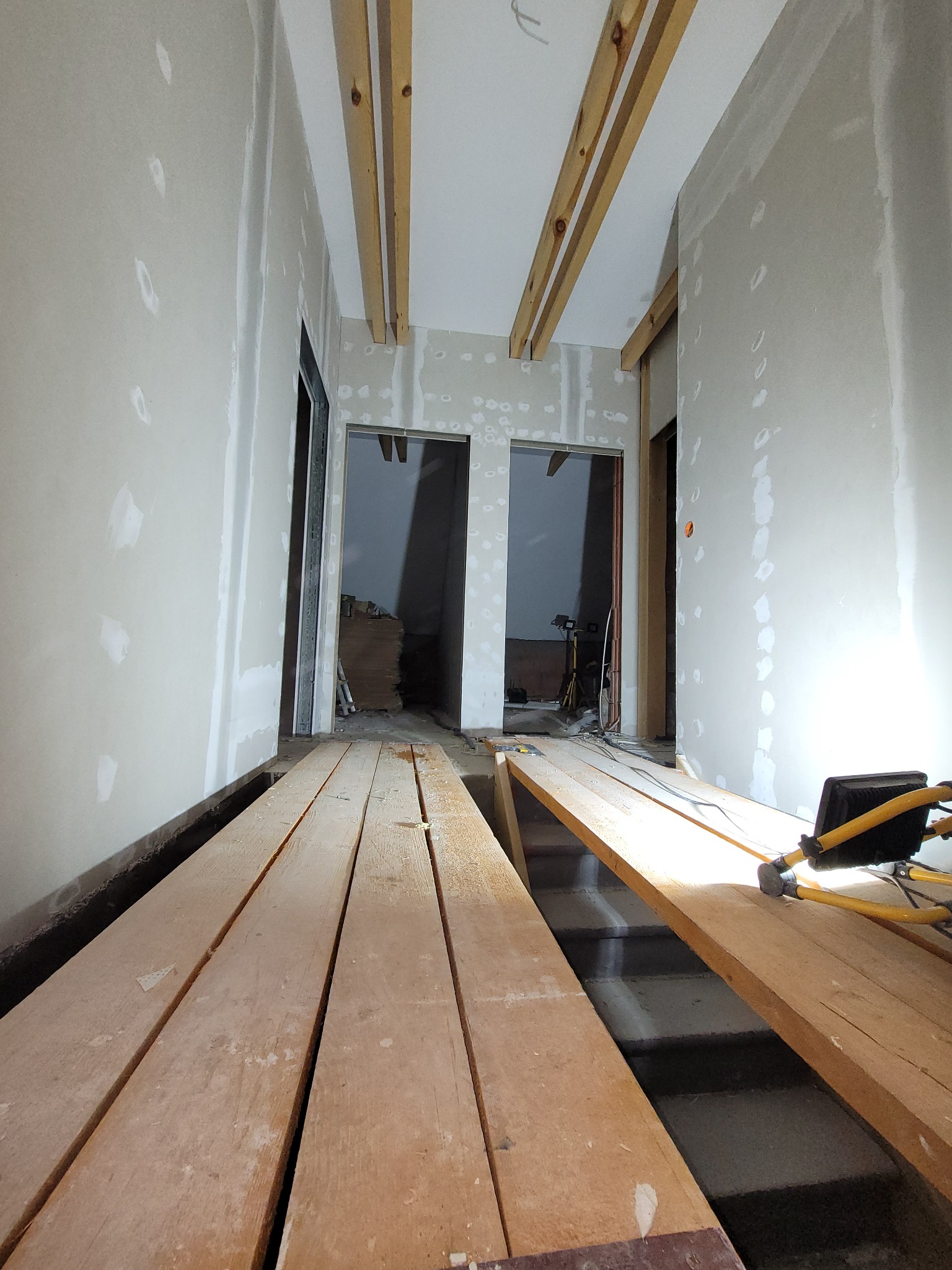 Wnętrze w trakcie remontu: ściany z płyt gipsowo-kartonowych z widocznymi łączeniami, drewniane belki pod sufitem, prowizoryczne podesty robocze i lampa budowlana.