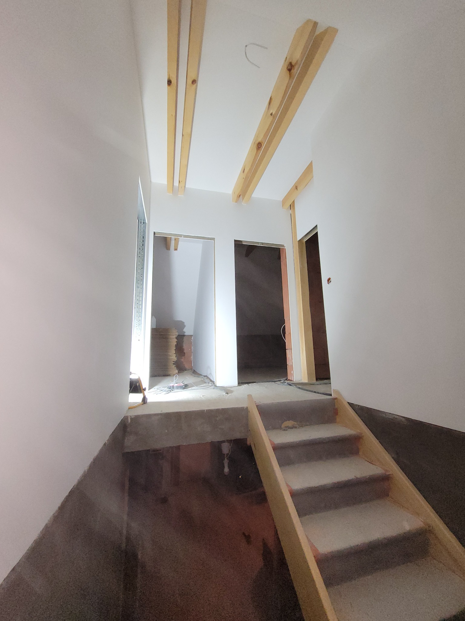 Wnętrze w trakcie remontu: surowe drewniane belki pod sufitem, schody z drewnianą konstrukcją, widoczne przygotowania do montażu ościeżnic drzwiowych, sterta płyt w głębi pomieszczenia.