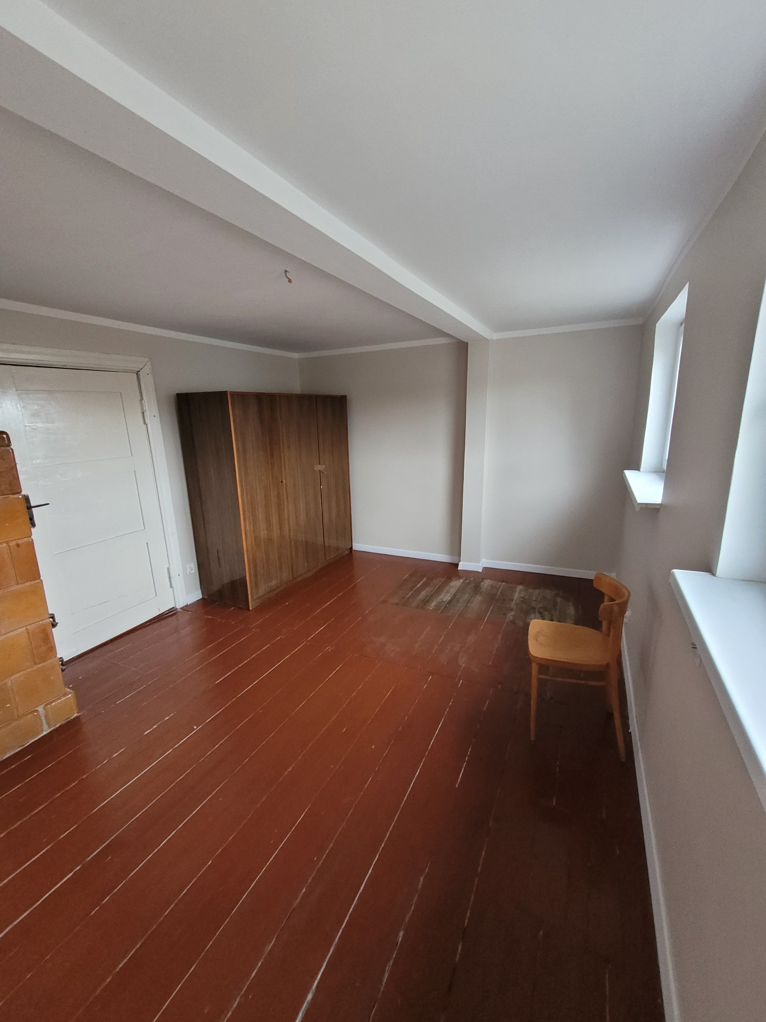 Wnętrze pokoju przed remontem: brązowa podłoga z widocznymi śladami użytkowania, ściany w odcieniach szarości, drewniana szafa, krzesło pod oknem i fragment pieca kaflowego przy drzwiach.