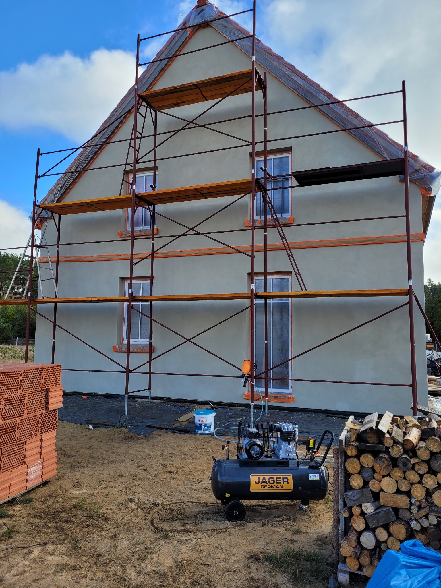 Dom w trakcie tynkowania elewacji, widoczne rusztowanie, stos cegieł, kompresor JAGER GERMAN i ułożone drewno opałowe.