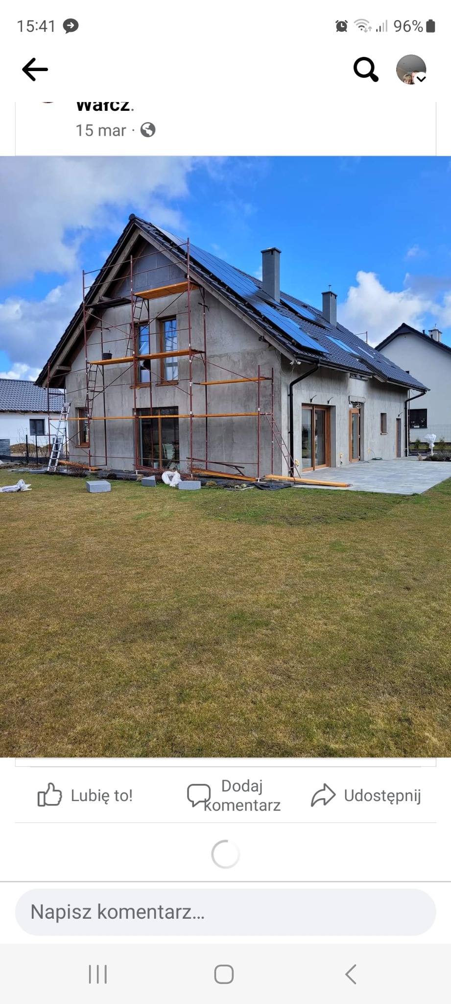 Dom jednorodzinny w trakcie prac elewacyjnych, widoczne rusztowania, szary tynk surowy, panele fotowoltaiczne na dachu, trawnik z przodu, częściowo wykończony taras z tyłu.