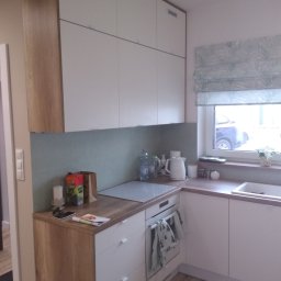 ARTISAN MEBLE SPÓŁKA Z OGRANICZONĄ ODPOWIEDZIALNOŚCIĄ - Nowoczesna kuchnia z białymi szafkami i drewnianymi akcentami, okap zintegrowany z szafką, roleta w liście przy oknie, na blacie czajnik i butelki z wodą.