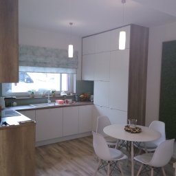 ARTISAN MEBLE SPÓŁKA Z OGRANICZONĄ ODPOWIEDZIALNOŚCIĄ - Nowoczesna kuchnia z białymi frontami szafek i drewnianymi wykończeniami, okrągły stół z białymi krzesłami, roleta w liście i panel z mchu na ścianie.