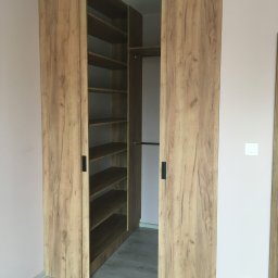 ARTISAN MEBLE SPÓŁKA Z OGRANICZONĄ ODPOWIEDZIALNOŚCIĄ - Otwarta szafa wnękowa z przesuwnymi drzwiami w odcieniach drewna, z widocznymi półkami i drążkiem na ubrania, na tle jasnoróżowej ściany.