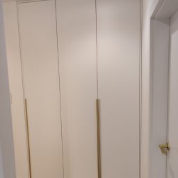 ARTISAN MEBLE SPÓŁKA Z OGRANICZONĄ ODPOWIEDZIALNOŚCIĄ - Biała, trzydrzwiowa szafa wnękowa z podłużnymi, złotymi uchwytami, widok z boku, z fragmentem drzwi wejściowych po prawej stronie.