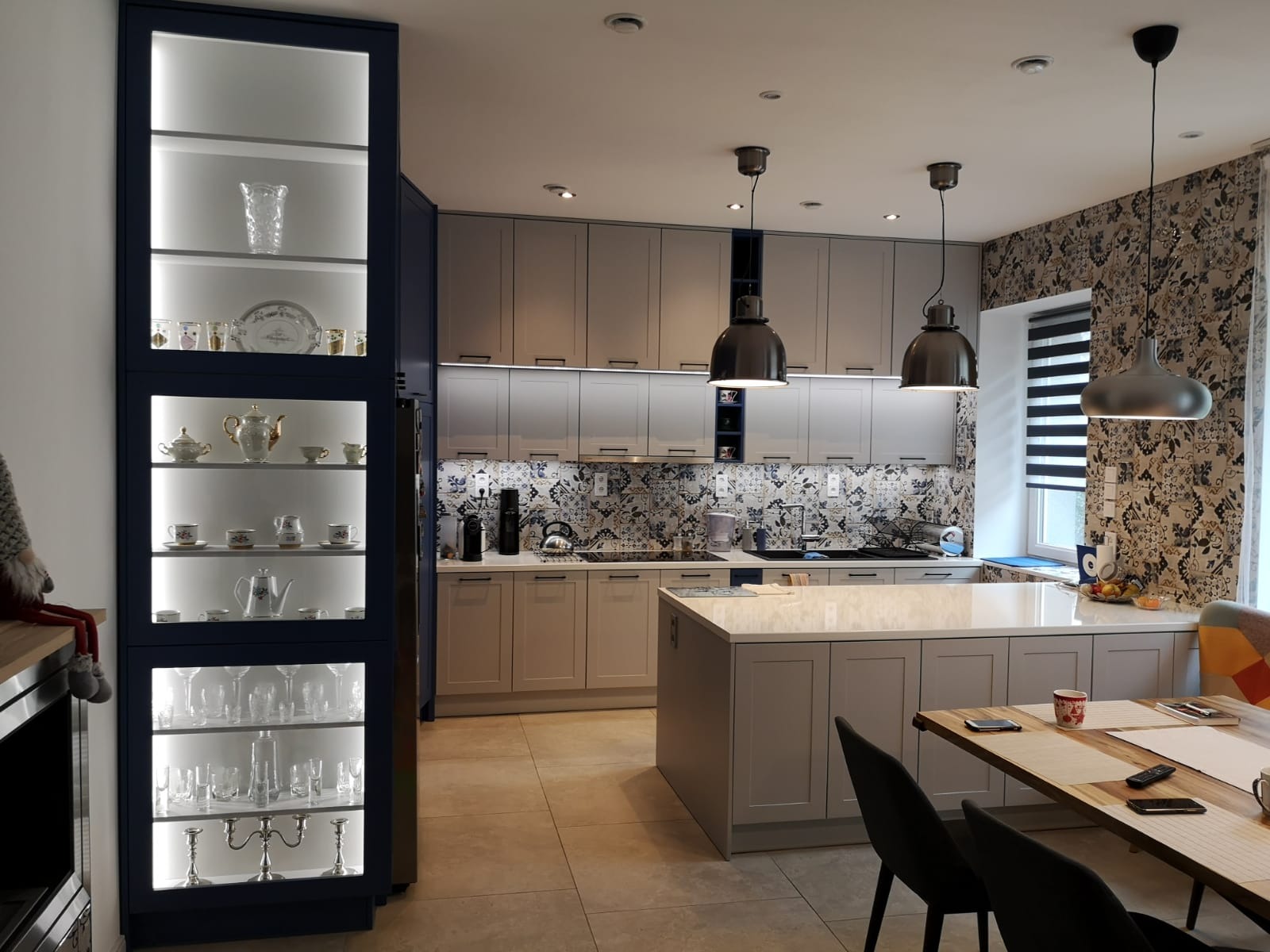 Jasnoszara kuchnia z wyspą, oświetlona szafka ekspozycyjna z porcelaną i szkłem, wzorzysta tapeta i trzy wiszące lampy.