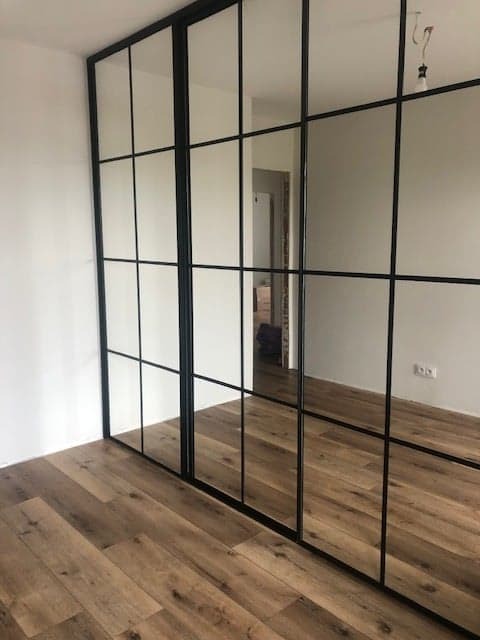 Nowoczesna szafa wnękowa z lustrzanymi drzwiami i czarnymi szprosami, widok na drewnianą podłogę i fragment wnętrza pokoju.