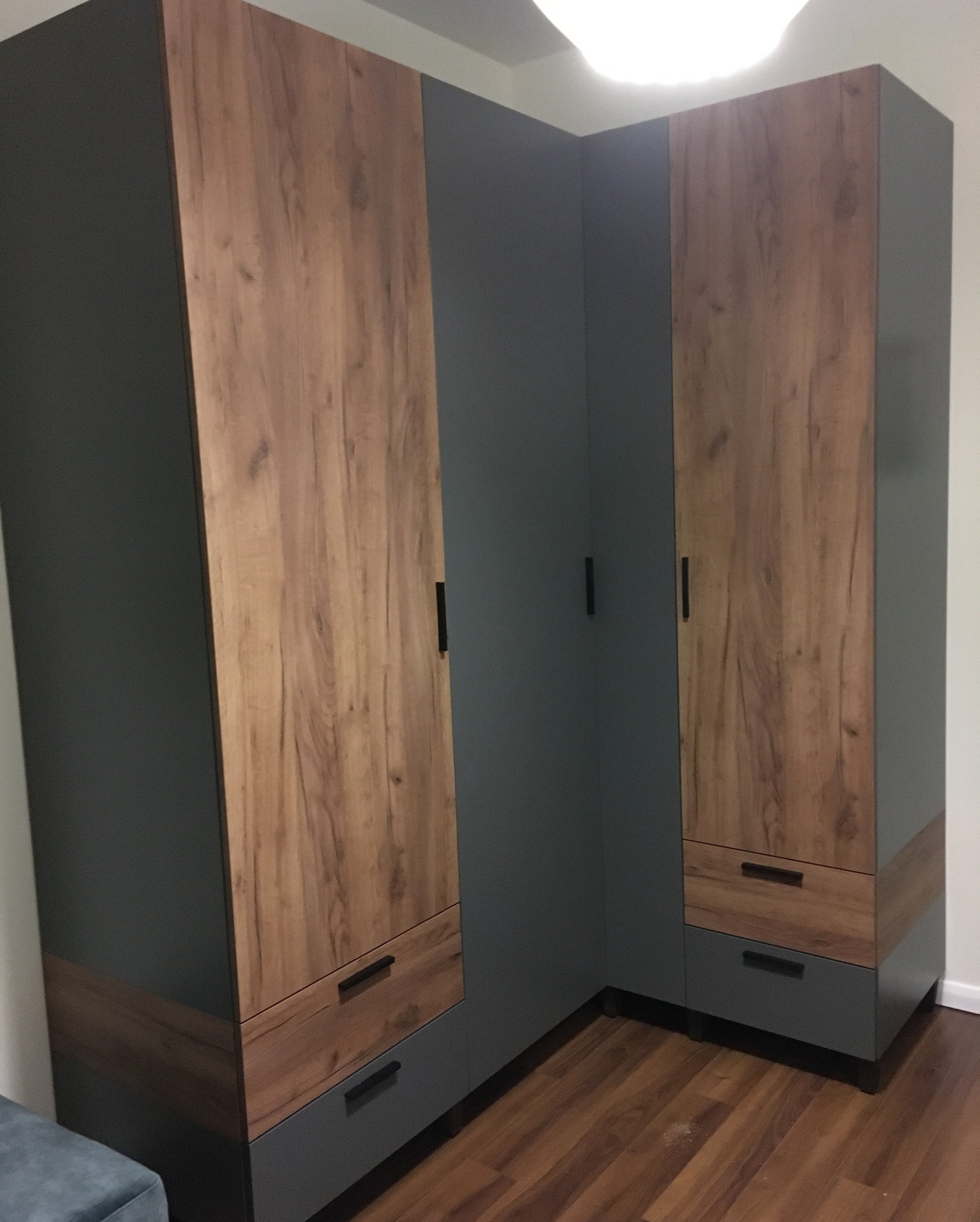 Narożna szafa z szarymi bokami i frontami z okleiną drewnopodobną, z czarnymi uchwytami, na parkiecie.