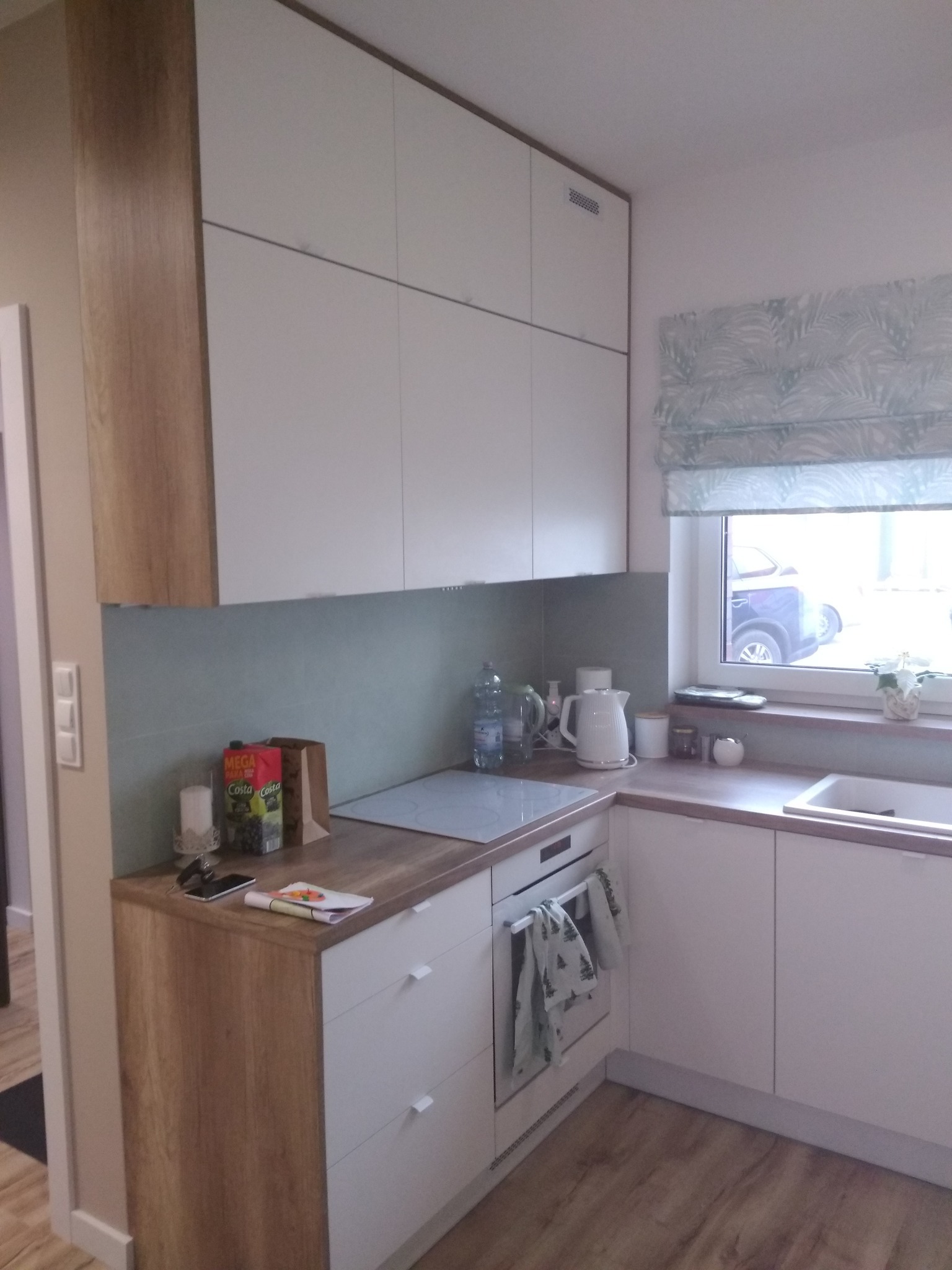 Nowoczesna kuchnia z białymi szafkami i drewnianymi akcentami, okap zintegrowany z szafką, roleta w liście przy oknie, na blacie czajnik i butelki z wodą.