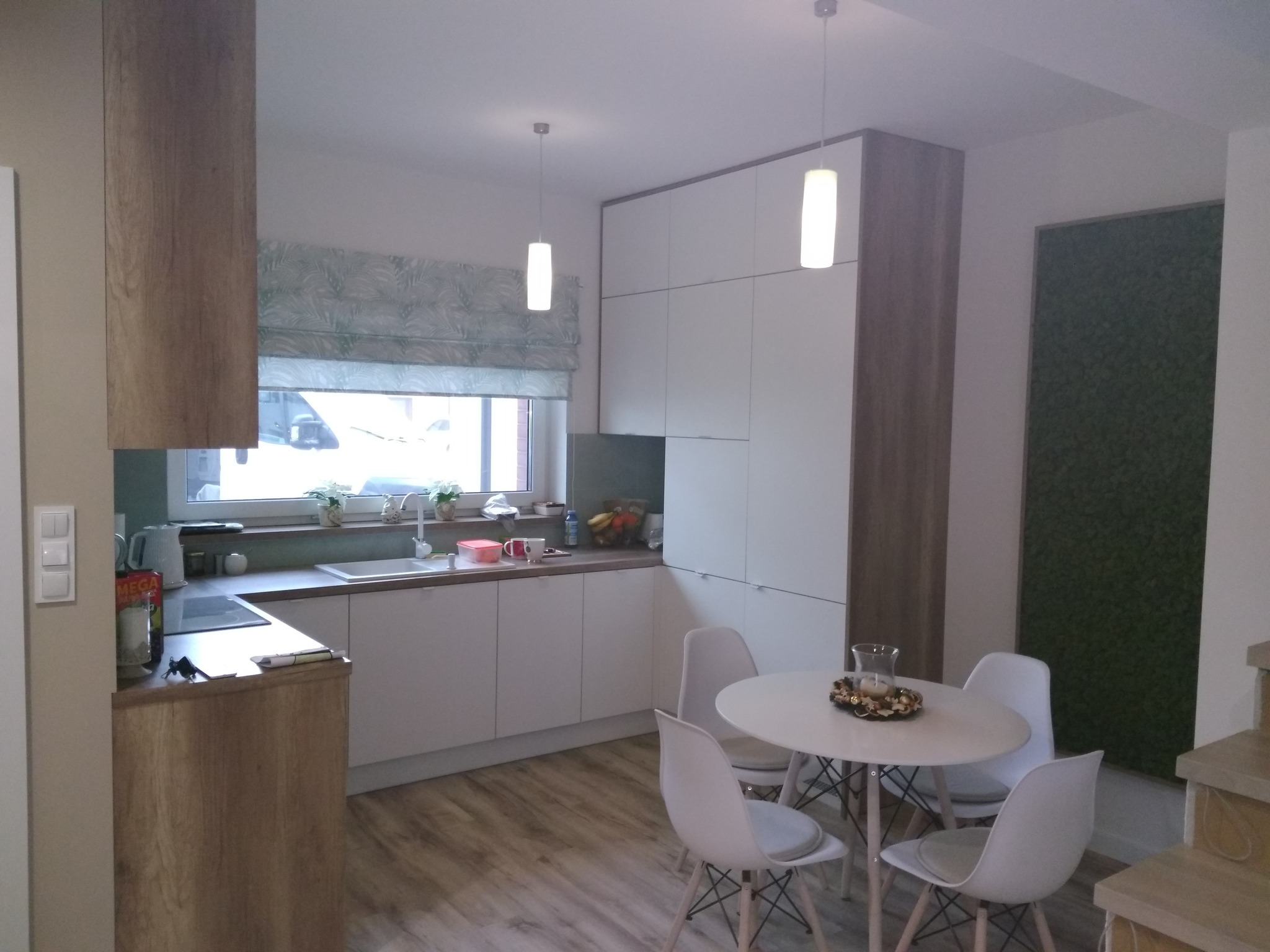 Nowoczesna kuchnia z białymi frontami szafek i drewnianymi wykończeniami, okrągły stół z białymi krzesłami, roleta w liście i panel z mchu na ścianie.