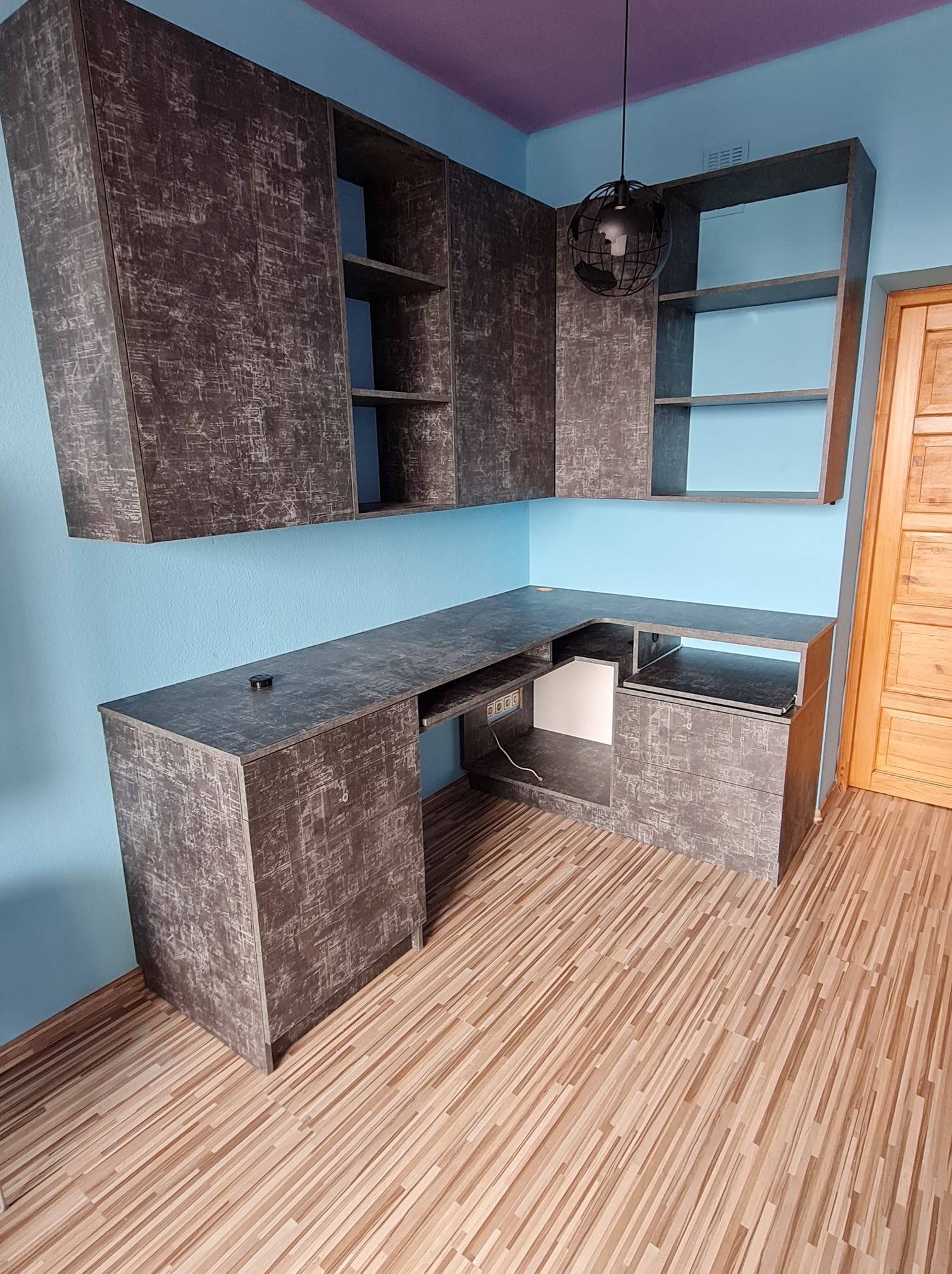 Kącik biurowy z szarymi szafkami wiszącymi i biurkiem narożnym z blatem o teksturze betonu, oświetlony lampą w kształcie kuli, na tle błękitnej ściany i drewnianej podłogi.