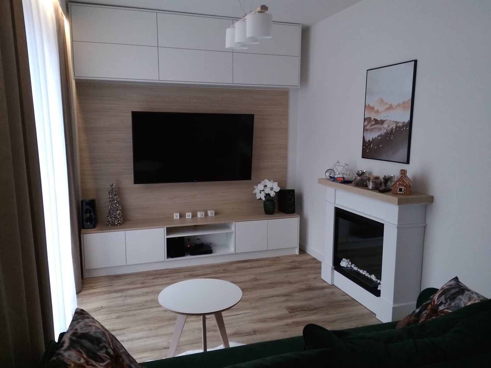 Nowoczesny salon z zabudową meblową pod telewizor, szafkami wiszącymi, kominkiem elektrycznym i drewnianą podłogą; dekoracje świąteczne w tle.