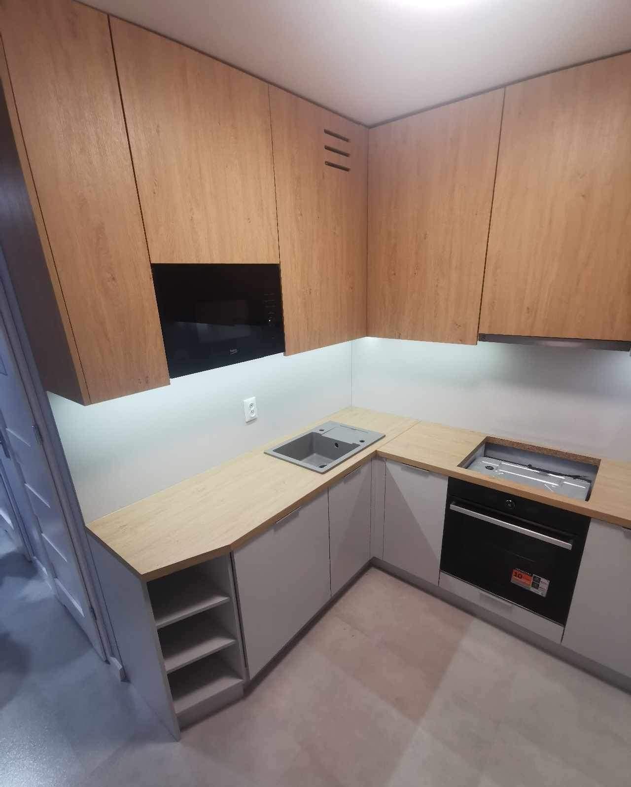 Nowoczesna kuchnia narożna z drewnianymi górnymi szafkami i szarymi dolnymi, z wbudowanym piekarnikiem i szarym zlewozmywakiem, oświetlona listwą LED pod szafkami.