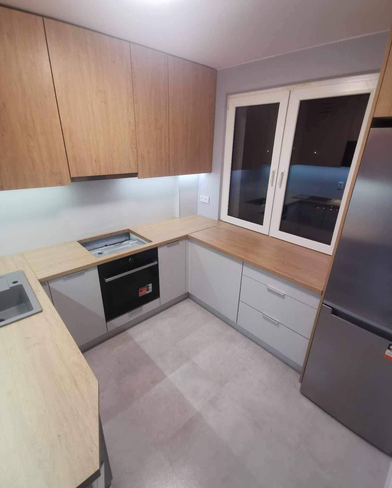 Jasna kuchnia z drewnianymi górnymi szafkami, szarymi dolnymi szafkami i blatem z jasnego drewna przy oknie, widoczny zlew i piekarnik.