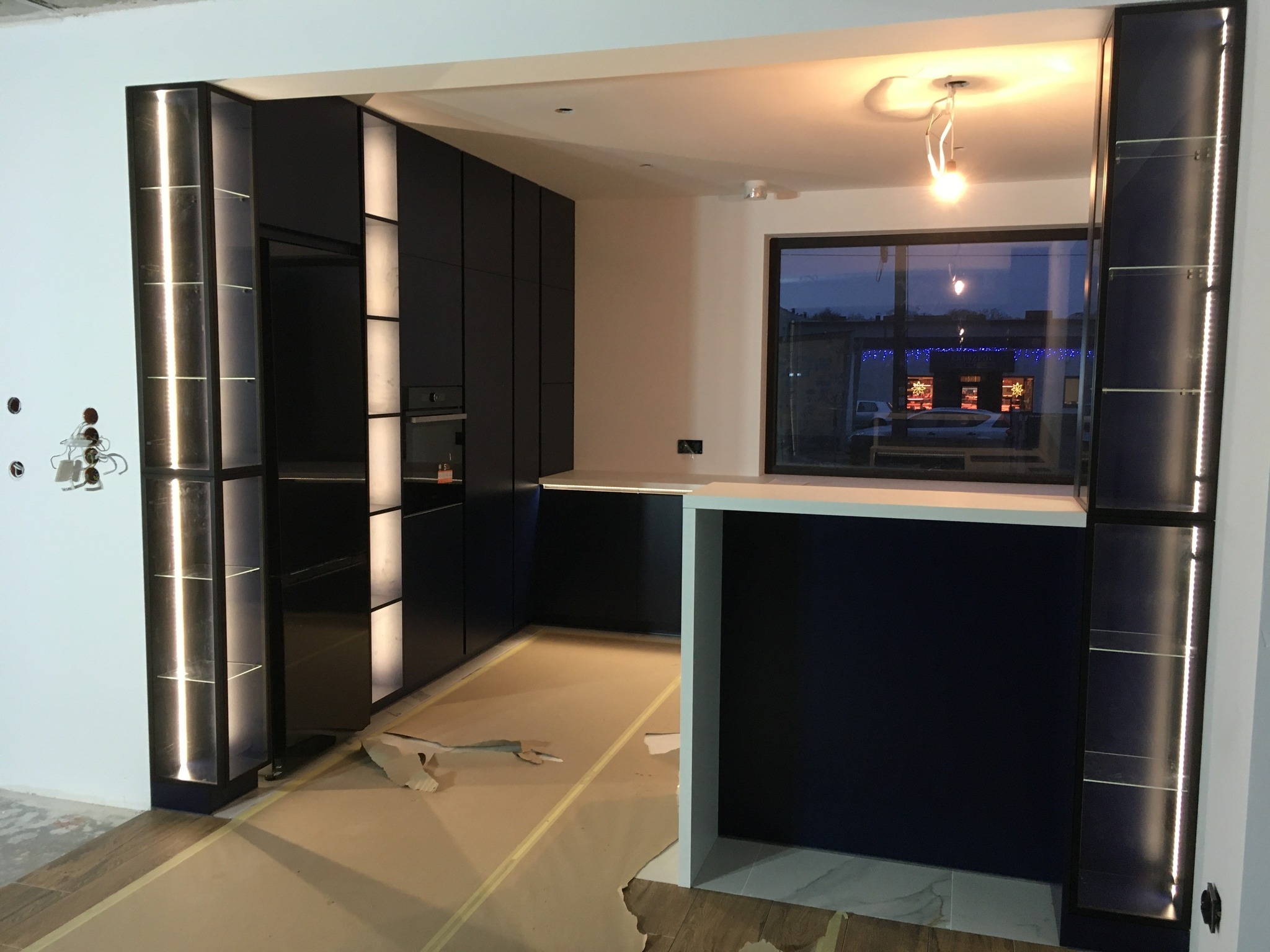 Ciemna zabudowa kuchenna z podświetlanymi witrynami, widok na okno z odbijającymi się światłami, podłoga zabezpieczona folią malarską.