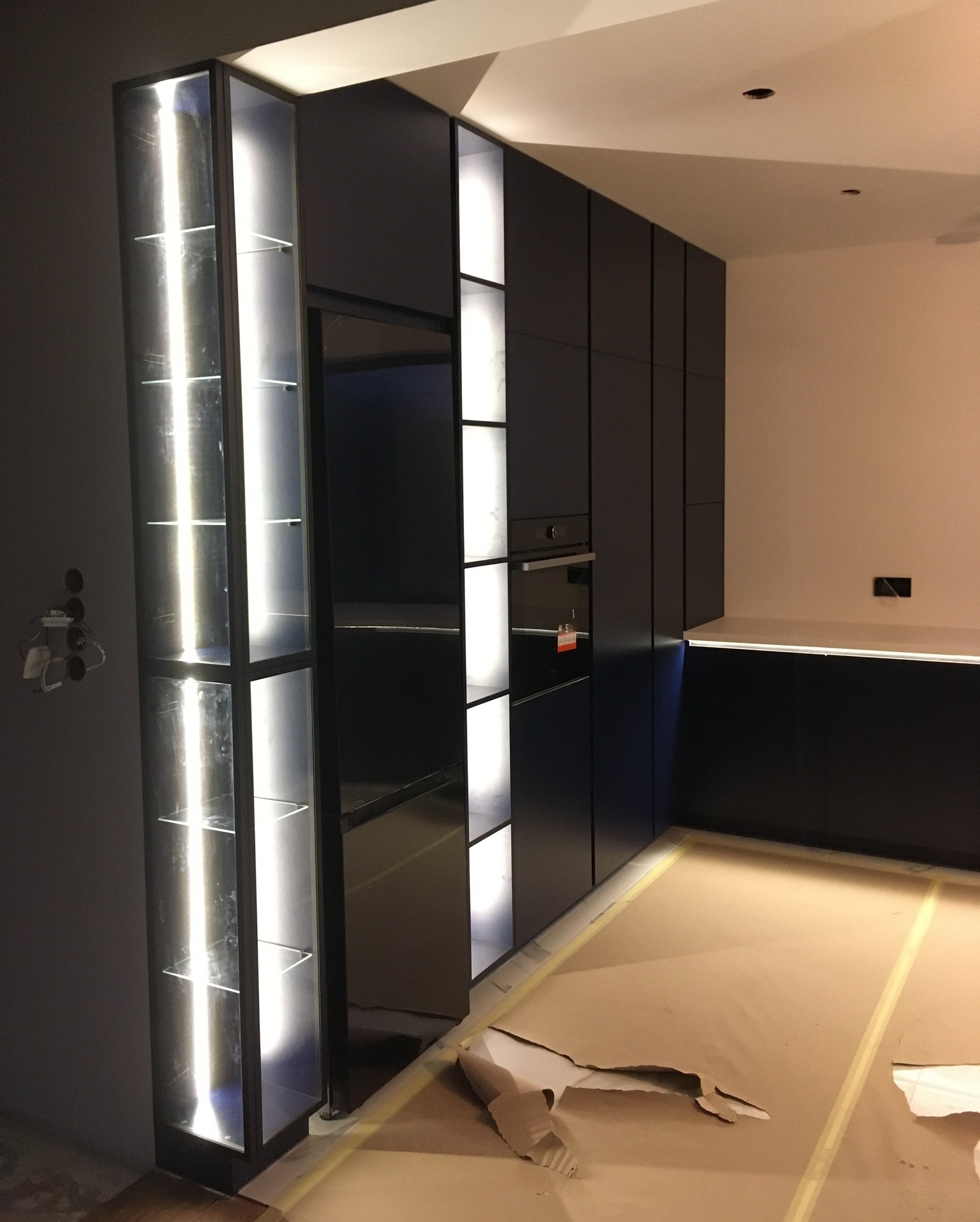 Ciemna zabudowa kuchenna z podświetlanymi półkami, widoczny piekarnik i zabezpieczona podłoga.