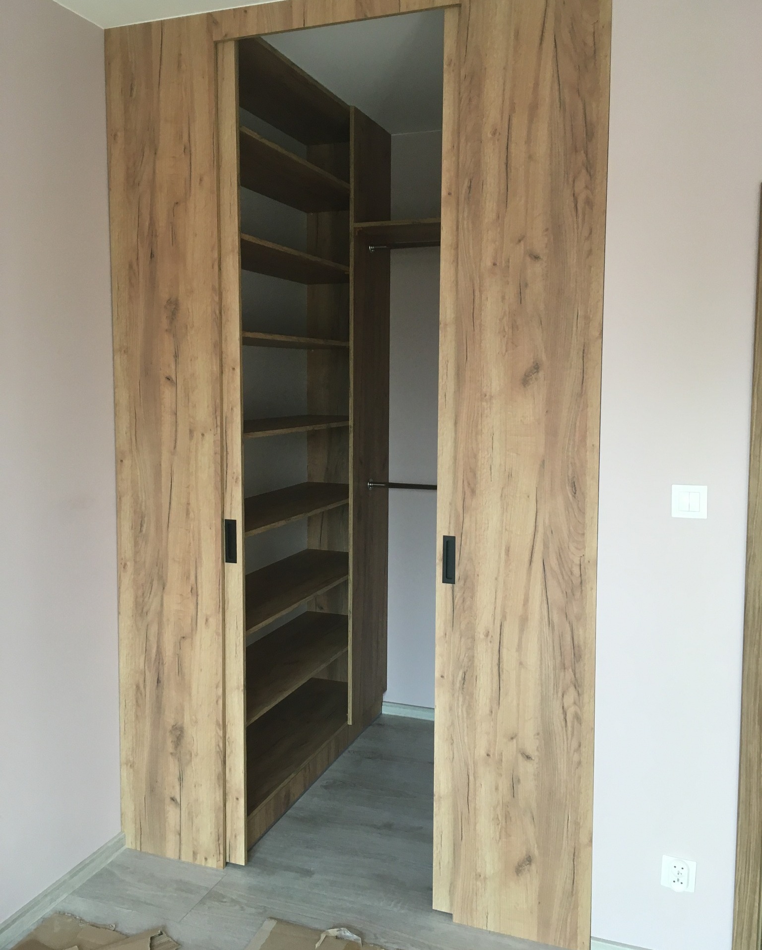 Otwarta szafa wnękowa z przesuwnymi drzwiami w odcieniach drewna, z widocznymi półkami i drążkiem na ubrania, na tle jasnoróżowej ściany.