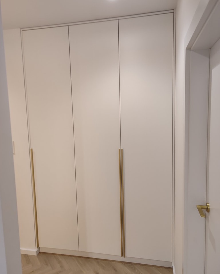 Biała, trzydrzwiowa szafa wnękowa z podłużnymi, złotymi uchwytami, widok z boku, z fragmentem drzwi wejściowych po prawej stronie.
