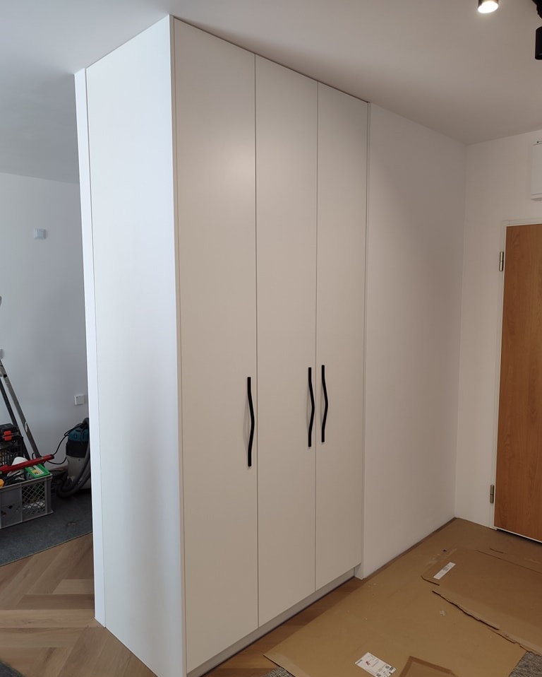 Biała, czterodrzwiowa szafa wnękowa z minimalistycznymi czarnymi uchwytami, widoczna na tle parkietu i fragmentu kartonów zabezpieczających podłogę podczas montażu.