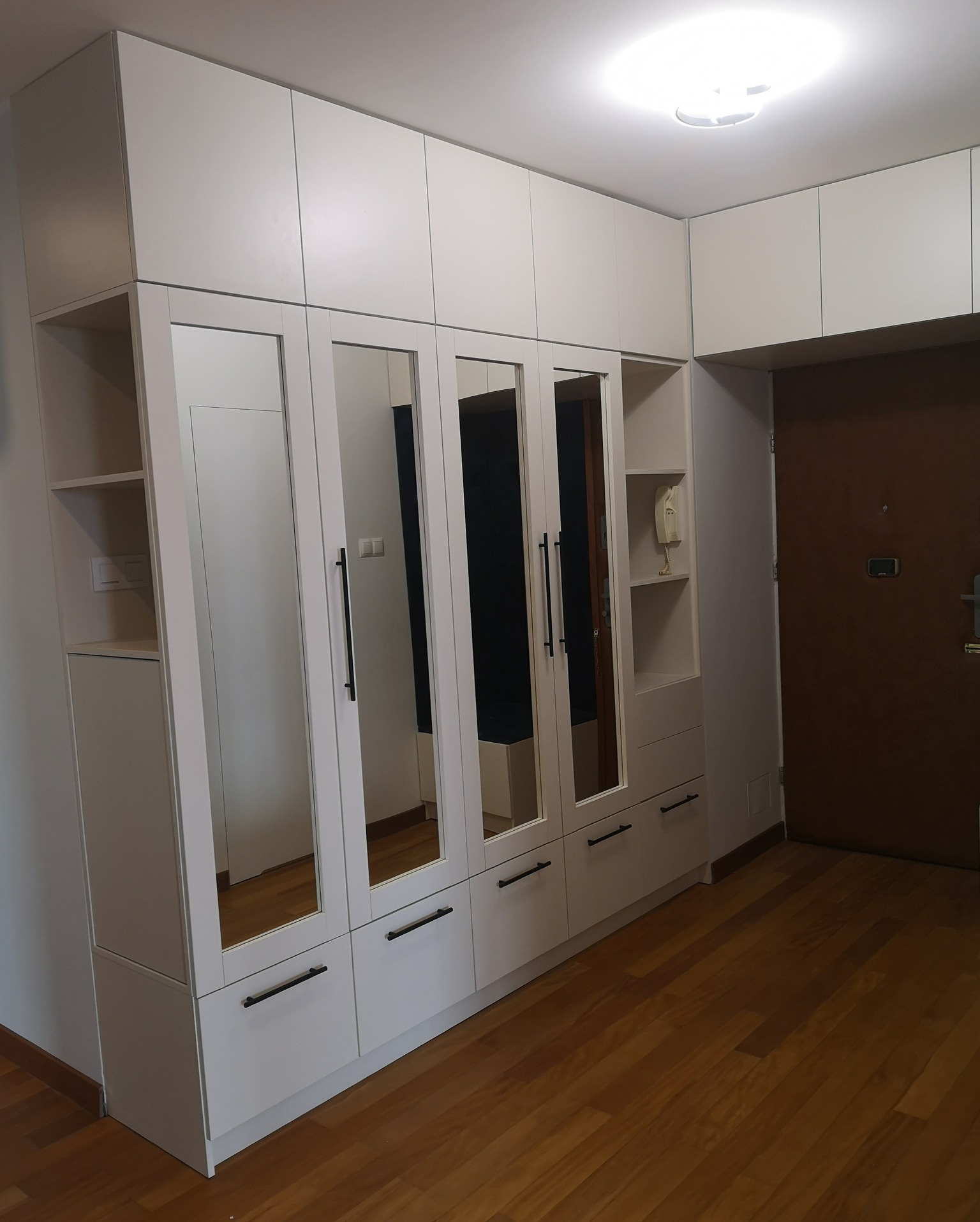 Biała szafa wnękowa z lustrami na frontach, z czarnymi, minimalistycznymi uchwytami, zabudowana od podłogi do sufitu, z dodatkowymi półkami po bokach, widok na drewnianą podłogę i drzwi wejściowe...