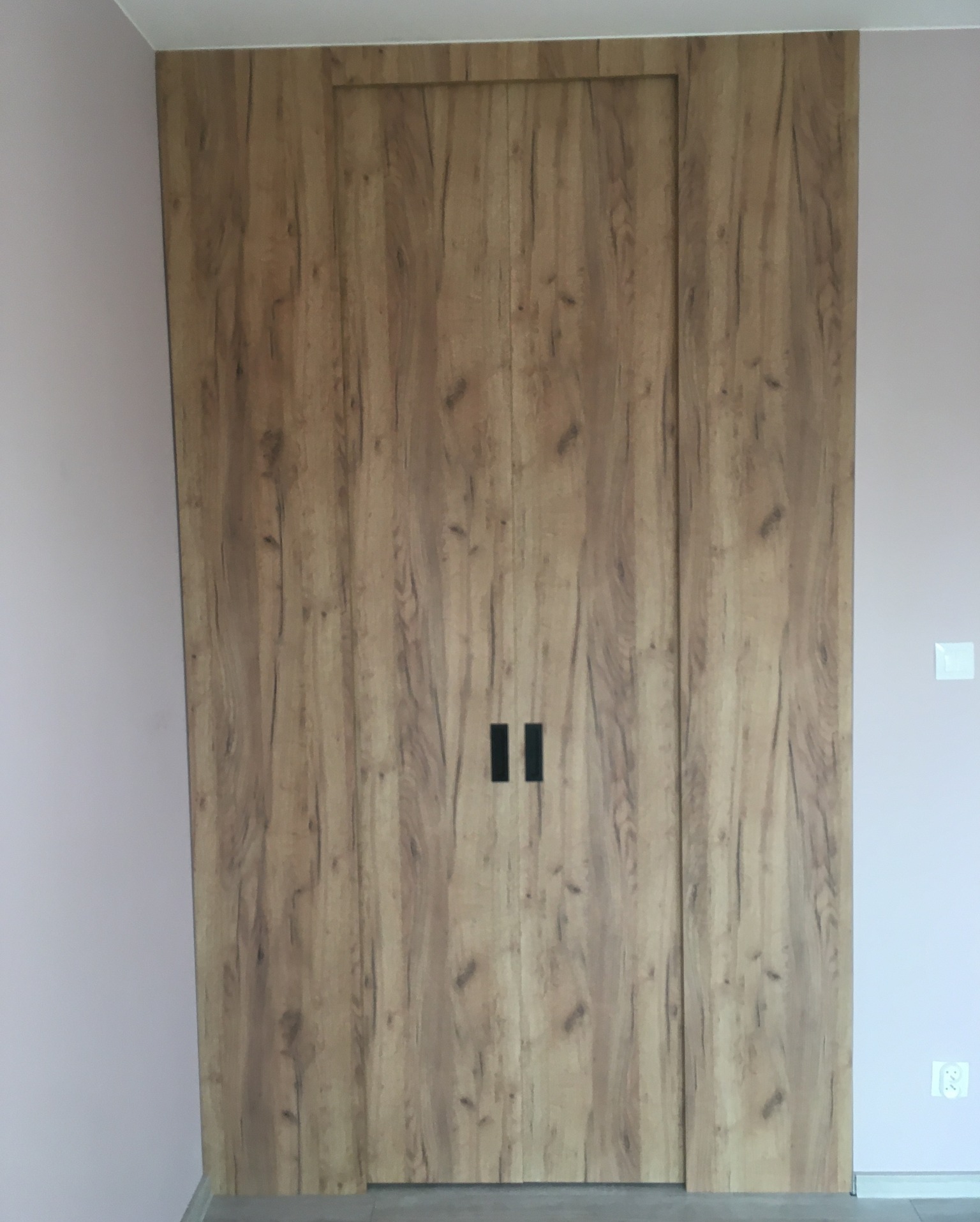 Zamknięte przesuwne drzwi szafy wnękowej z okładziną imitującą jasne drewno, z minimalistycznymi czarnymi uchwytami, na tle jasnoróżowej ściany.