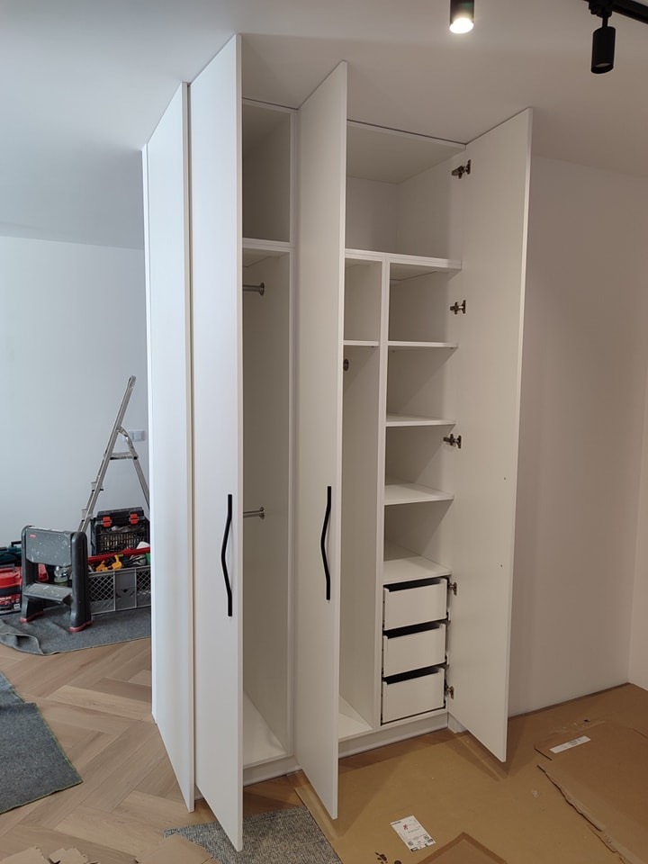 Biała, trzydrzwiowa szafa z otwartymi drzwiami, widoczne półki, drążek na ubrania i trzy szuflady w dolnej części, czarne, falowane uchwyty, w tle drabina i narzędzia, podłoga zabezpieczona kartonem.