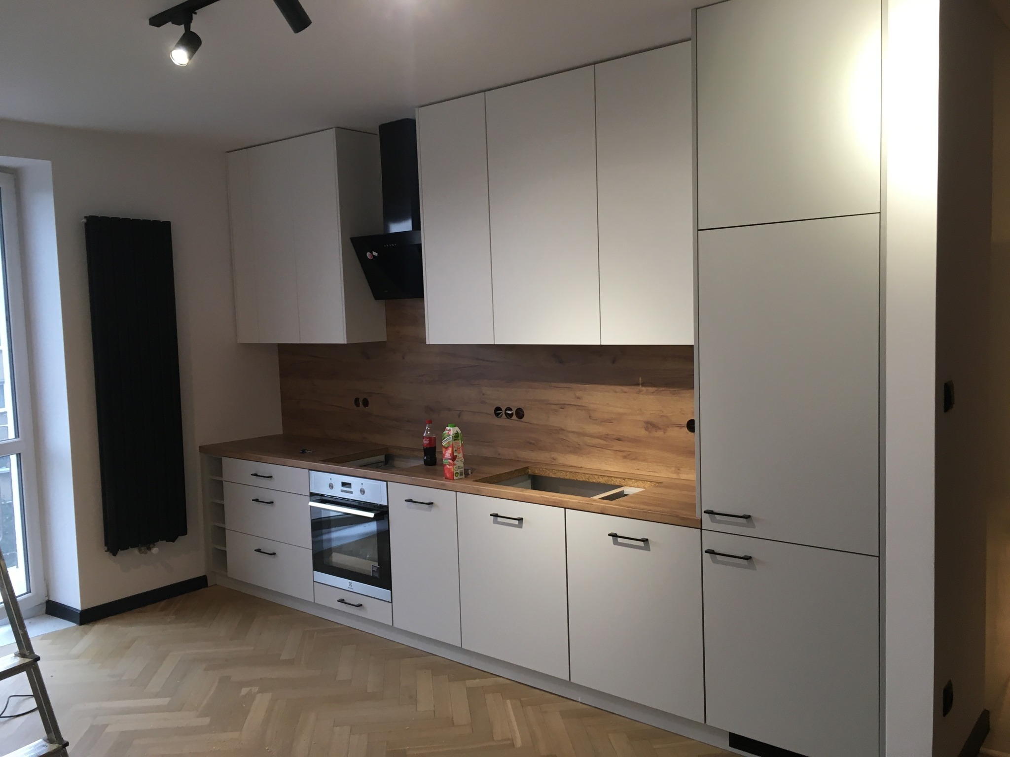 Nowoczesna kuchnia z białymi frontami szafek i drewnianym blatem, widoczne niedokończone podłączenia elektryczne i brak baterii przy zlewie, podłoga w jodełkę, drabina oparta o ścianę przy oknie.