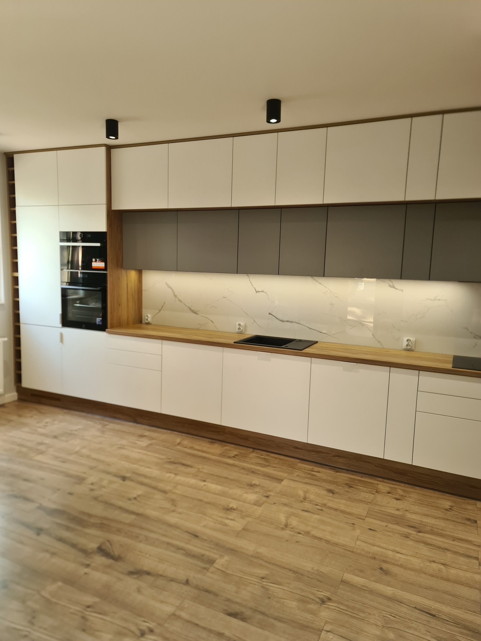 Nowoczesna kuchnia z białymi i szarymi szafkami, drewnianym blatem i podłogą, oświetlona punktowymi lampami sufitowymi.