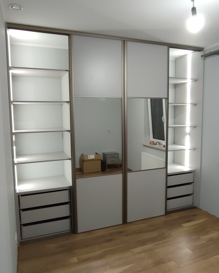 Biała szafa wnękowa z przesuwnymi drzwiami, z lustrem na jednym skrzydle, oświetleniem LED wewnątrz i szufladami, widoczna na tle drewnianej podłogi.