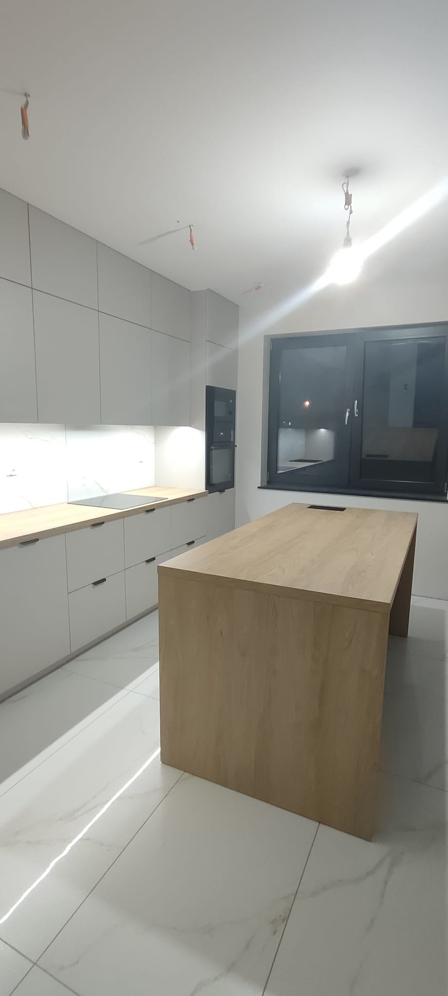 Nowoczesna kuchnia z szarymi szafkami, drewnianym blatem i wyspą kuchenną z jasnego drewna, oświetlona listwą LED pod szafkami wiszącymi, na podłodze białe płytki imitujące marmur.