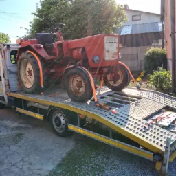 Czerwony, starszy traktor zabezpieczony pomarańczowymi pasami na platformie lawety, widok z boku, częściowo zacieniony.