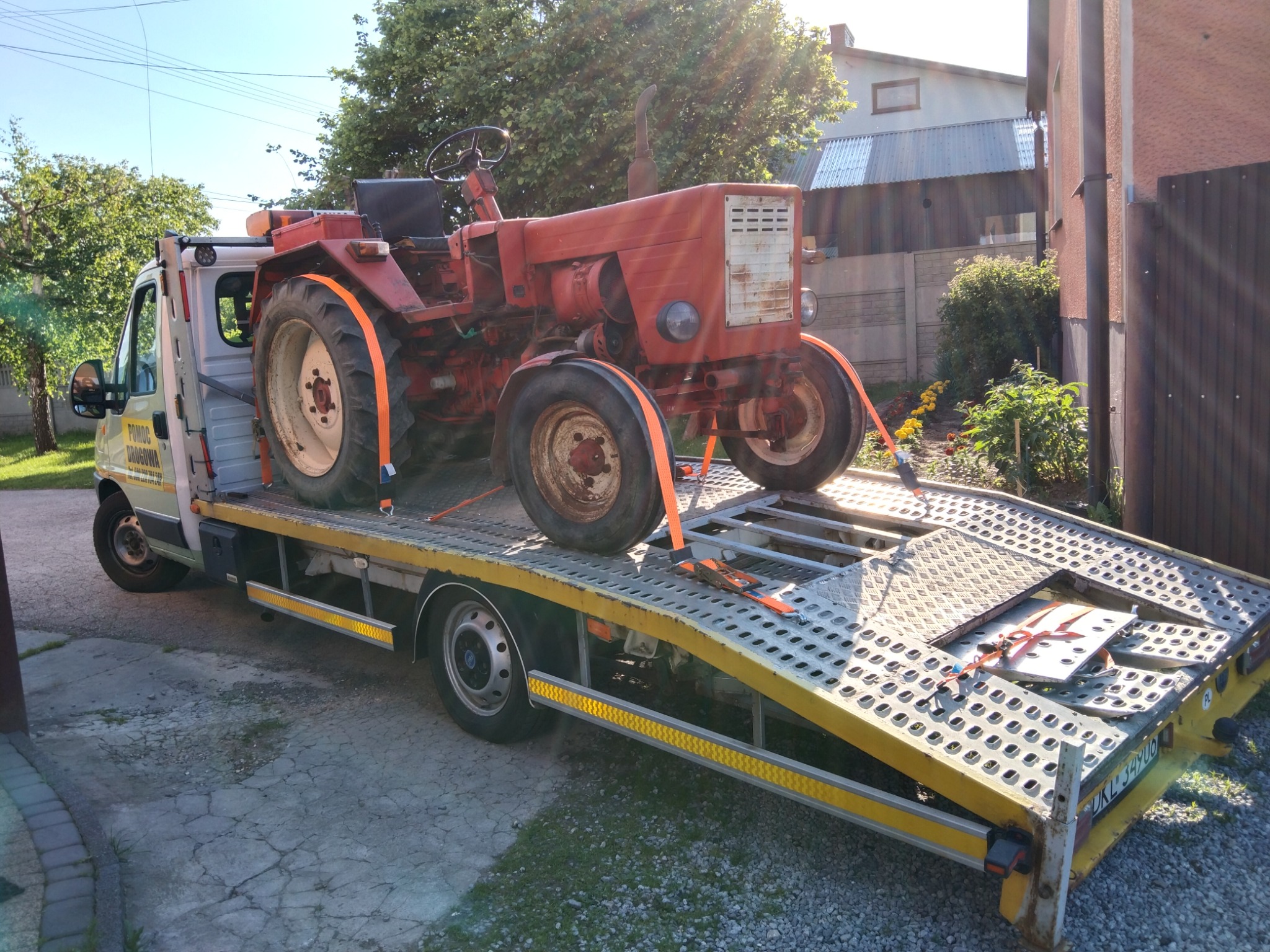 Czerwony, starszy traktor zabezpieczony pomarańczowymi pasami na platformie lawety, widok z boku, częściowo zacieniony.