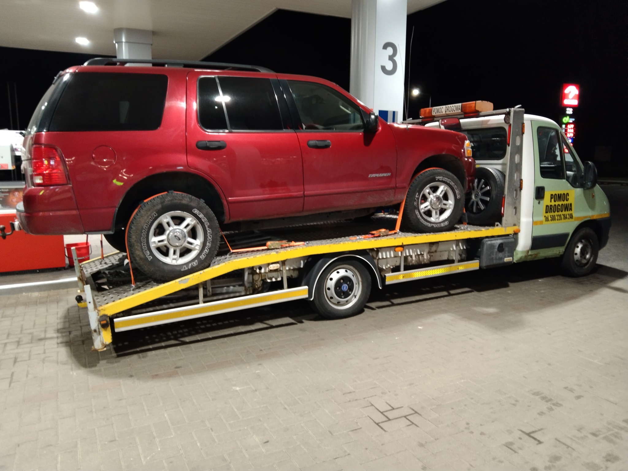 Czerwony SUV załadowany na żółtą lawetę pomocy drogowej na stacji benzynowej nocą. Na aucie pomocy drogowej widoczny napis 'POMOC DROGOWA' i numer telefonu.