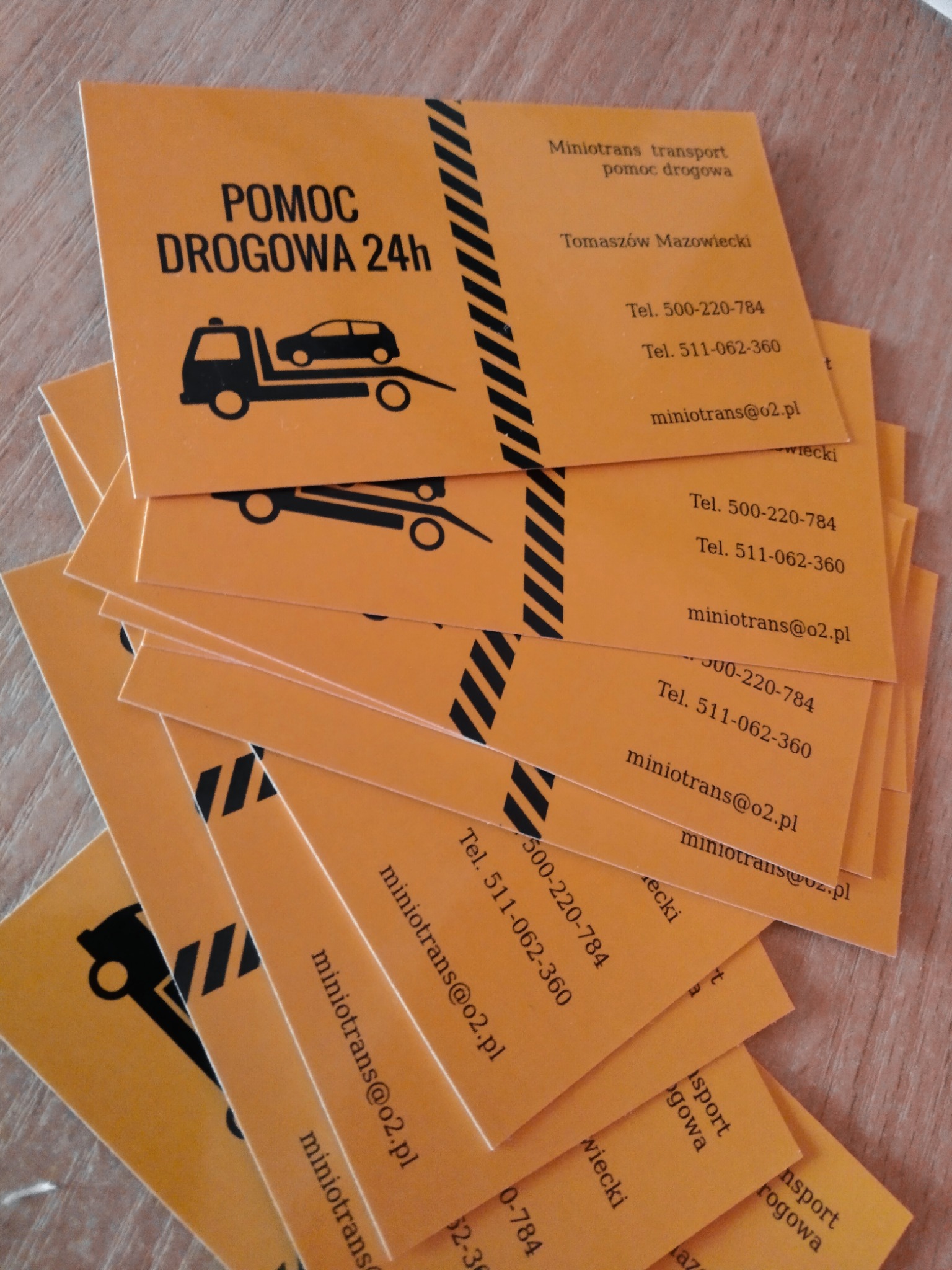 Stos wizytówek firmy Minotrans transport pomoc drogowa, w kolorze pomarańczowym z czarnym nadrukiem, zawierających logo lawety z samochodem oraz dane kontaktowe.