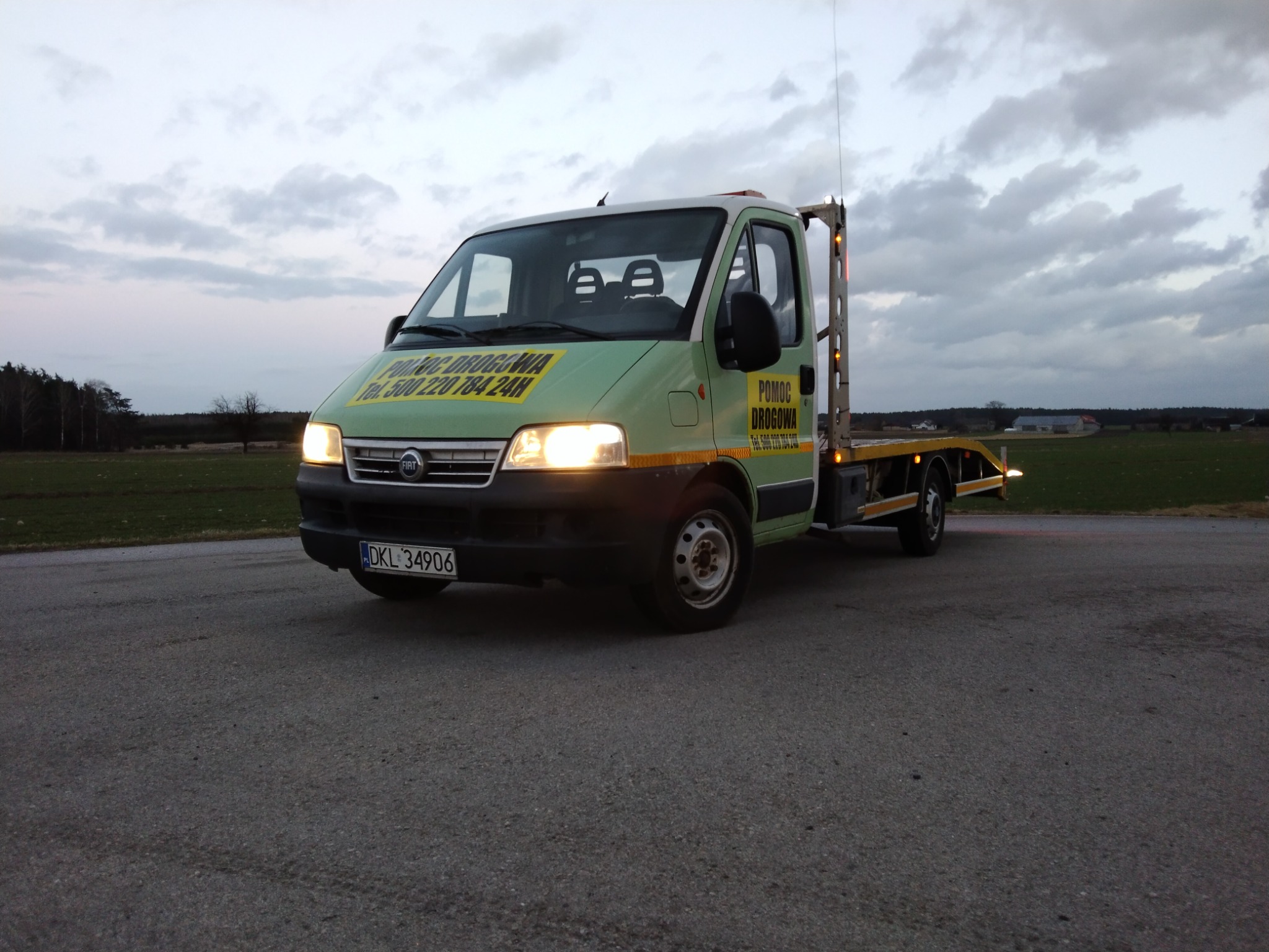 Zielona laweta Fiat z żółtym napisem 'POMOC DROGOWA' i numerem telefonu, stojąca na asfaltowej drodze na tle pola i zachmurzonego nieba o zmierzchu.