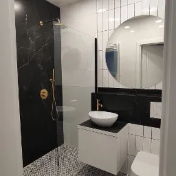 Nowoczesna łazienka z czarno-białą marmurową ścianą prysznicową, złotą armaturą, umywalką nablatową i geometryczną podłogą.