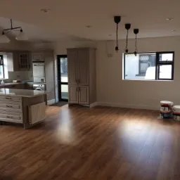 Jasne, przestronne wnętrze z nowymi panelami podłogowymi, świeżo pomalowanymi ścianami i farbami w wiadrach przy ścianie, gotowe do dalszych prac wykończeniowych.