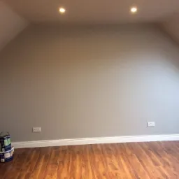 Pomalowane na szaro poddasze ze skośnymi ścianami, widoczne puszki z farbą przy ścianie i parkiet na podłodze.