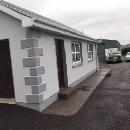 Odnowiona elewacja domu jednorodzinnego z dekoracyjnymi elementami imitującymi kamień, szare ściany i białe okna, widok z boku na tle pochmurnego nieba.