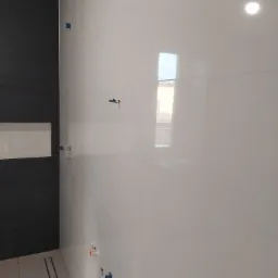 Ściana łazienki wyłożona białymi, błyszczącymi płytkami z widocznymi instalacjami wodnymi i elektrycznymi, czarna ściana z wnęką, odpływ liniowy w podłodze.