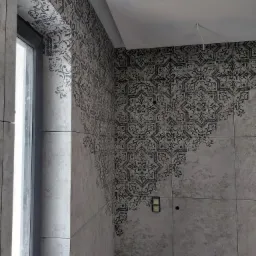 Ściana łazienki wyłożona płytkami z wzorem dekoracyjnym, częściowo pokrywającym szarą powierzchnię, widoczny fragment okna i wystający kabel ze ściany.