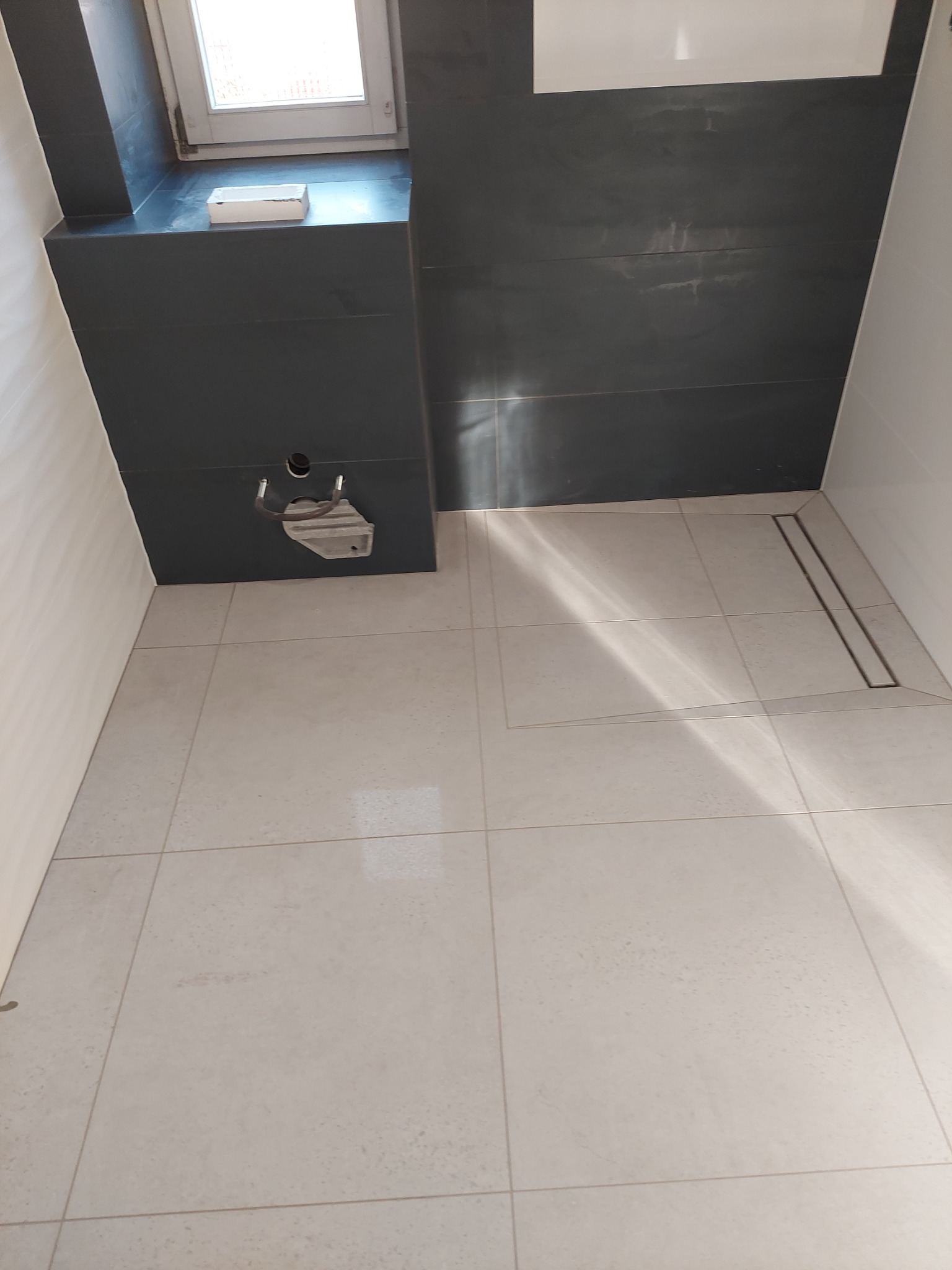 Wnętrze łazienki w Ostrowcu Świętokrzyskim w trakcie prac wykończeniowych, widoczne ułożone jasne płytki podłogowe, czarne płytki ścienne i odpływ liniowy w podłodze.