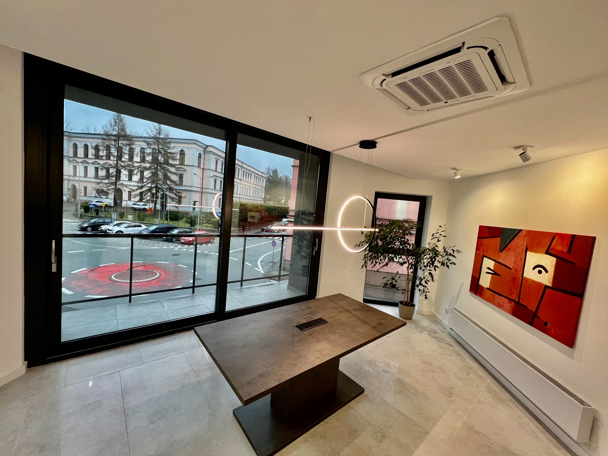 Nowoczesne biuro z dużym oknem, przez które widać budynek sądu, minimalistyczny stół konferencyjny i okrągła lampa sufitowa.