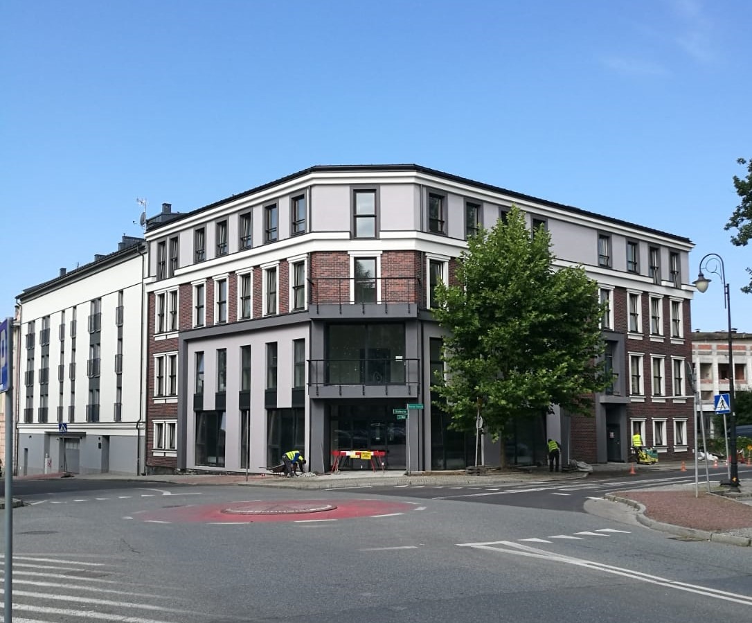 Nowoczesny, trzypiętrowy budynek biurowy z elementami cegły i szarej elewacji, widoczni pracownicy w kamizelkach odblaskowych wykonujący prace brukarskie na chodniku przed budynkiem.