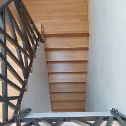 Widok z góry na jasne, drewniane schody z czarną, geometryczną balustradą, przylegające do jasnej ściany.