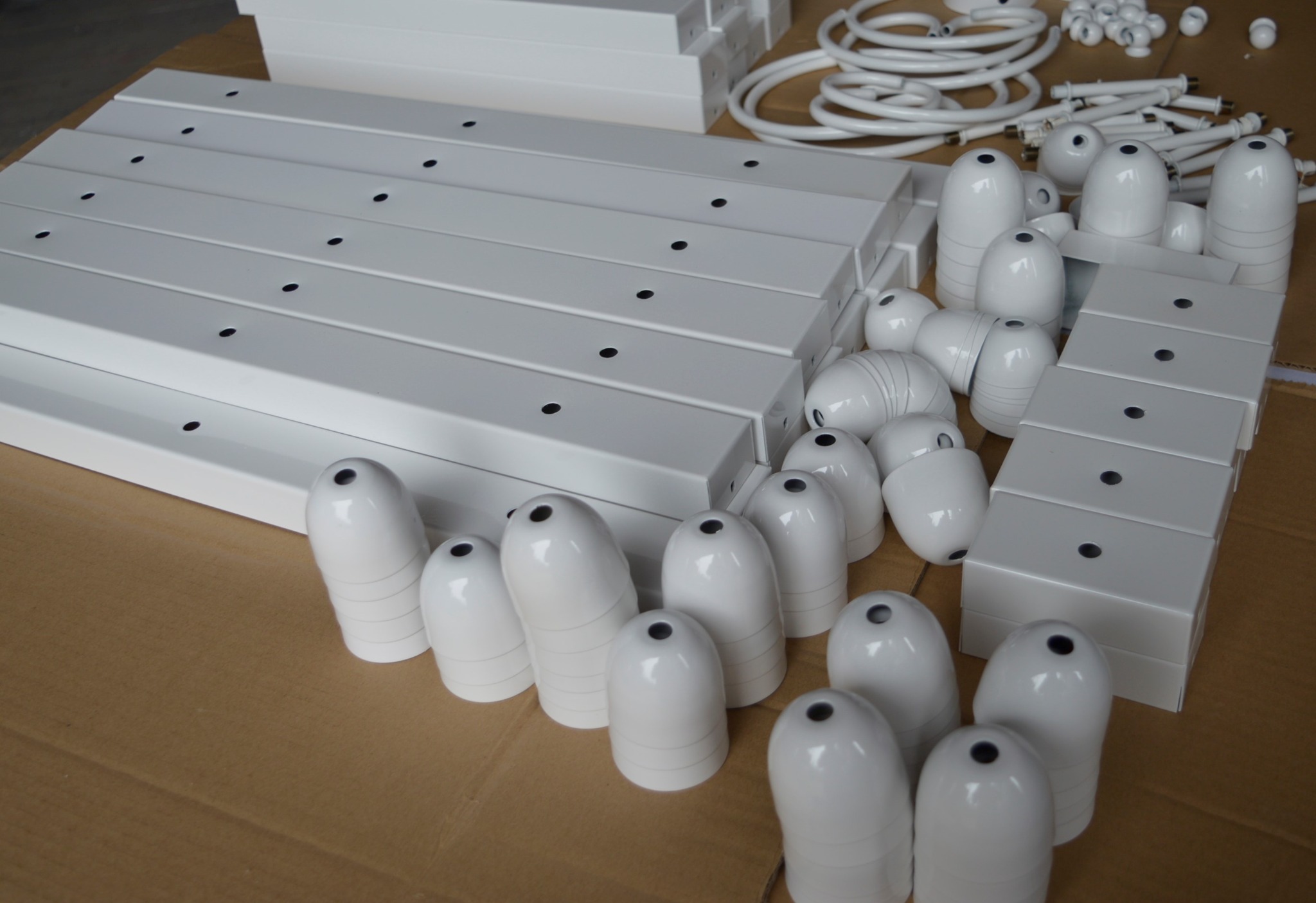 Białe, metalowe elementy oświetleniowe, w tym prostokątne obudowy z otworami i klosze w kształcie łezki, ułożone na kartonowej powierzchni.