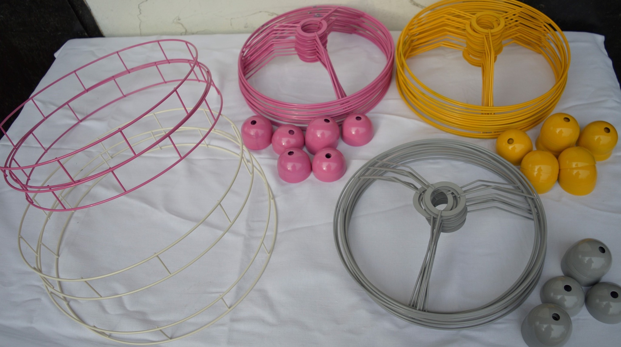 Metalowe stelaże do lamp i kuliste osłonki w kolorach: kremowym, różowym, żółtym i szarym, ułożone na białym obrusie.