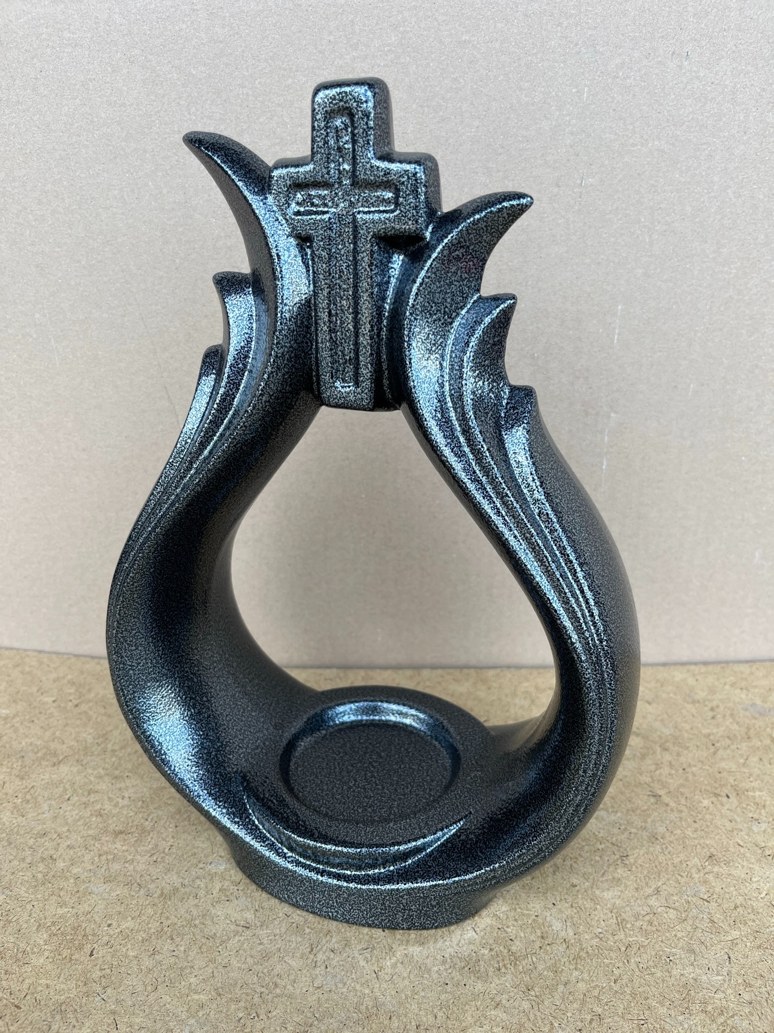 Metalowa, czarna, błyszcząca ozdoba z krzyżem na górze, na brązowym tle.